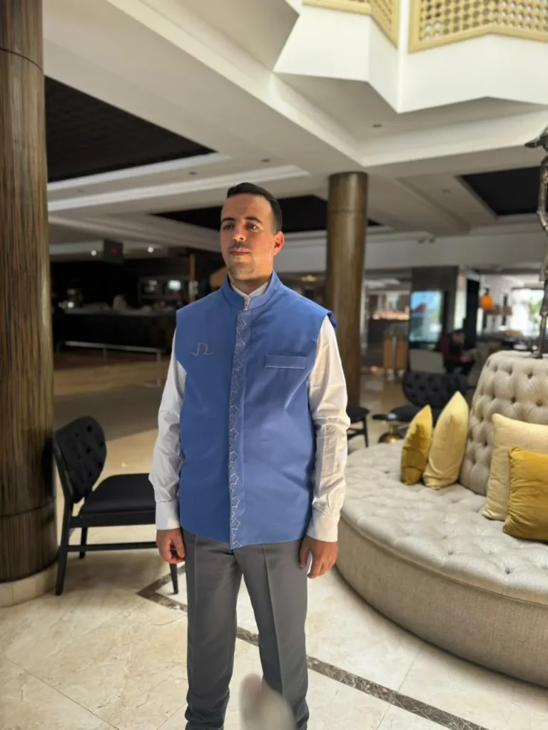 Employé d’hôtel dans le lobby en gilet sans manches bleu à col mao et broderie traditionnelle, chemise blanche et pantalon gris